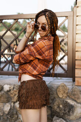 a girl with a dreadlocked hairstyle poses in summer outdoor, bright sunlight, dressed in a plaid...