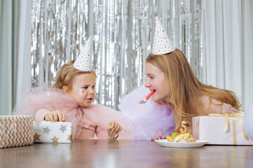 Obraz na płótnie Canvas Children's emotions. 5-year-old girl in party hat and festive dress is upset about something at her birthday celebration, her older sister is trying to make her laugh and calm her down.
