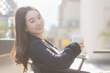 Business beautiful Asian  female in a dark blue suit is sitting on a chair and smiling happily. In front of the coffee shop in the city on a sunny morning.