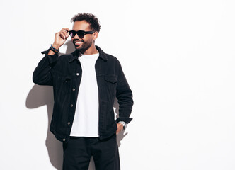 Handsome smiling hipster model. Sexy unshaven man dressed in black  summer shirt and jeans clothes. Fashion male posing near white wall in studio at sunny day. Isolated. In sunglasses