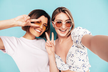 Two young beautiful smiling brunette hipster female in trendy summer  clothes. Sexy carefree women posing blue yellow wall. Positive models having fun. Cheerful and happy.Taking Pov selfie