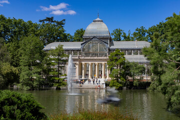Fototapeta na wymiar Crystal Palace. Building located in the Retiro Park in Madrid with its glass windows surrounded by trees and green vegetation on a sunny day and some clouds in the sky, in Spain. Europe. Photography.