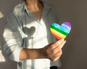 female hands hold a LGBT heart and a sunbeam creates a heart-shaped shadow on the chest,
lgbt pride, rainbow colored heart, LGBT symbol