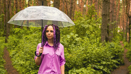 A woman with an umbrella in her hand in the rain. A girl in a purple shirt under an umbrella in the woods. A woman with dreadlocks stands in a park with an umbrella
