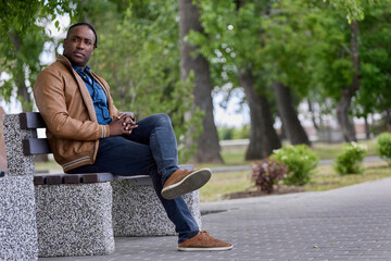 Obraz premium Handsome black man sits on a bench in the park waiting for another person. An African-American man sat down on a bench in an alley in a park, resting from a long road.