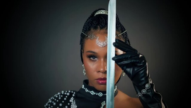 closeup portrait african fantasy woman warrior queen holding sword in hands strokes touches sharp blade, sexy looks at camera. Beauty face black dress girl princess tiara moon, dark gray studio 