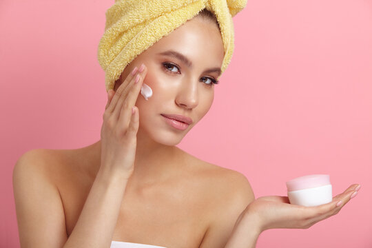 Seductive female model applies cream on cheek at pink wall