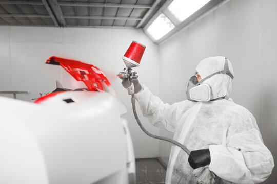 Male worker sprays red paint with spray gun on car bumper part in car maintenance service paint room.