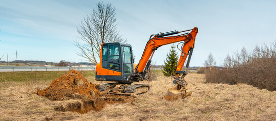 Banner image.Mini excavator digging earth in a field or forest near roadway.Digging trenches for a gas pipeline or oil pipeline.Spring,autumn blue sky background. © ARVD73