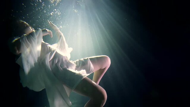 young woman is sinking in dark water of sea or lake, underwater view against sun rays from up