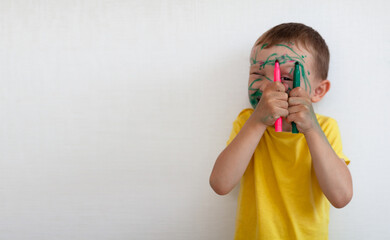 A 4-year-old boy painted his face with a marker. The boy stands against a white wall, holding...