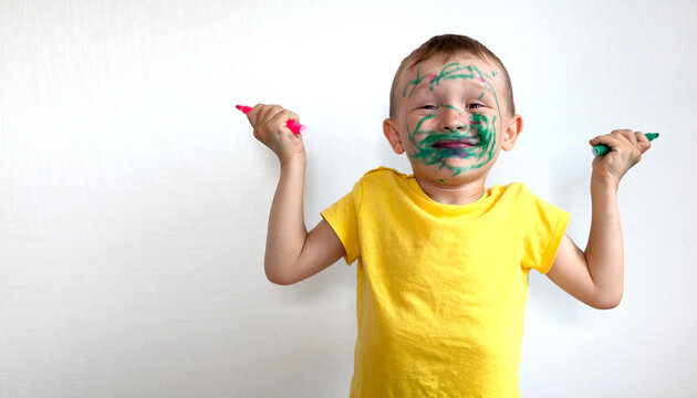 A 4-year-old Boy Painted His Face With A Marker. The Boy Stands Against A White Wall, Holding Markers In His Hands And Smiling. Banner With Space For Text