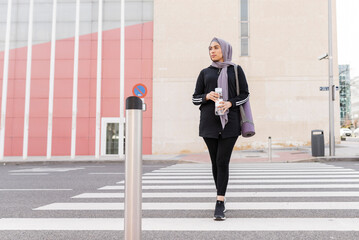 muslim woman in sportswear crossing street in the city, with bottle of water, a mat and backpack.