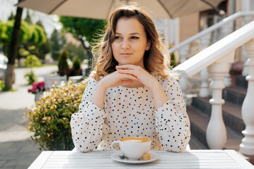 Portrait of stylish young woman with arms folded sitting at table with cup of coffee in street cafe on sunny day. Curly pretty lady resting in cafe, looking away outdoors.