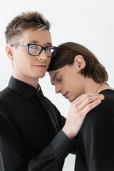 Smiling gay in eyeglasses looking at camera and hugging boyfriend isolated on grey.