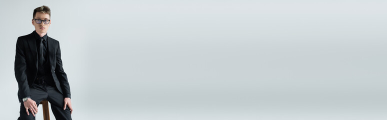 Young man in eyeglasses and suit sitting on chair isolated on grey, banner.