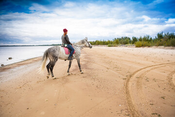 wearing jeans, jacket and warm hat girl is in the saddle on grey horse along seabeach