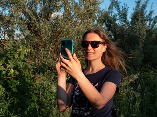 Happy young woman in sunglasses with smartphone calling friends talking walking outdoor summer sunny day. Millennial girl having video call using chat app technology. Authentic real people lifestyle.