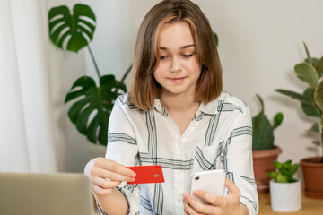 Female professional doing online payment through credit card while sitting at home office. Cheerful young girl shopping or banking online using mobile phone and credit card, living room interior.