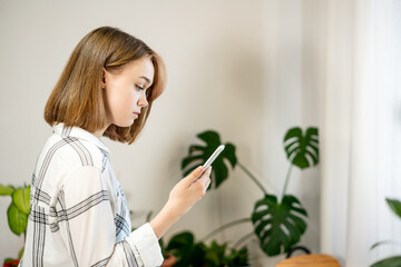 Young woman holding smartphone using mobile apps, chatting in social media at home in cozy living room with plants. Female entrepreneur working at home office. Remote work concept. A business woman is