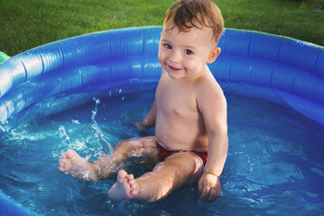 A one-year-old child bathes in a blue pool on a green fresh lawn. Boy bathes and splashes with water