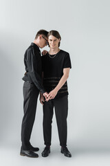 Full length of cheerful gay couple in black clothes holding hands on grey background.