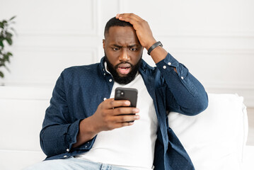 Oh no. Worried shocked young African-American bearded guy staring at smartphone screen, sitting on...