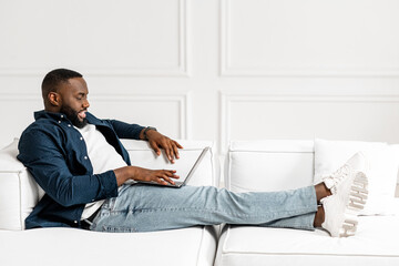 Full length side view African-American guy in casual shirt over t-shirt lying on the sofa with a laptop on his lap in bright modern apartment, black man resetting and chilling on the weekend at home