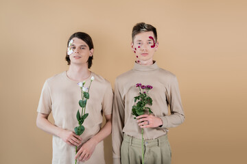 Same sex couple with petals on faces holding flowers isolated on beige.