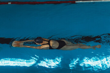 From above back view of professional woman in swimsuit swimming with foam board in water of indoors pool during training 