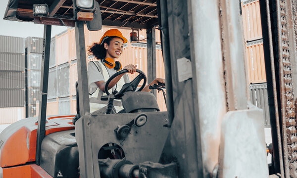 Female foreman with hard hat driving forklift at shipping container yard, portrait. Smiling multiracial industrial engineer woman in safety vest drives reach stacker to lift cargo box at logistic dock