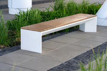 Empty concrete bench with wooden slats for sitting on tile among decorative grass and flowers in recreation area near modern office building. Garden landscape with chair in city park