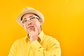Positive man in panama hat holding chin thinks about vacation, doubts, makes decision isolated on yellow studio background