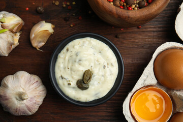 Creamy caper sauce in bowl and ingredients on wooden table, flat lay.
