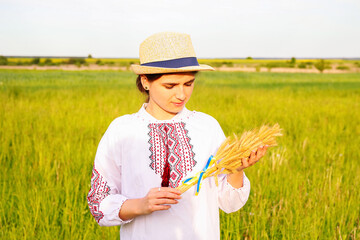 Defocus young woman in vyshyvanka and hat holding bouquet of ripe golden spikelets of wheat tied on...