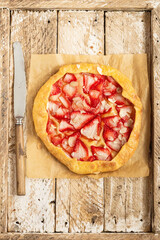 Galette with strawberry and almonds petals. Shortcrust pastry pie on the perchament paper. Galette in rustic style on the wooden tray. Summer pie with seasonal berries, knife with wooden handle