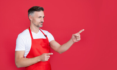 smiling mature houseman in apron on red background. pointing finger
