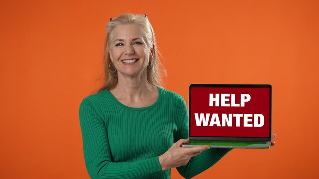 Smiling happy woman holding laptop with Help Wanted on screen on solid orange background.