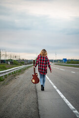 Woman with curly hair, in an unbuttoned plaid shirt, jeans, high heels, walking along the highway towards the horizon (with back to the viewer) with electric guitar. Hitchhiker or traveler musician