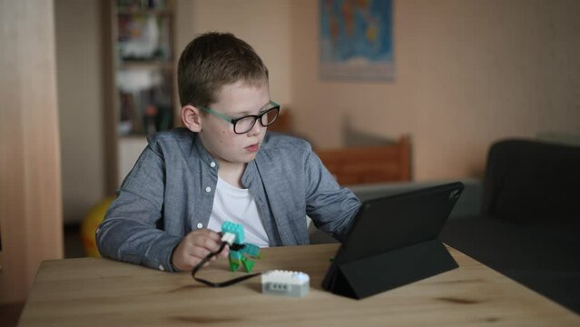 Caucasian boy in glasses sits at table, tries to program movement algorithm of toy from constructor on tablet. Child is surprised, embarrassed because of failed attempt, looks at screen in confusion.