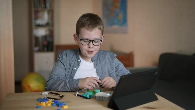 Child in glasses is dissatisfied, disassembles toy from constructor into parts and talks tensely. Boy is sitting at table in children room, there is tablet on the tabletop.