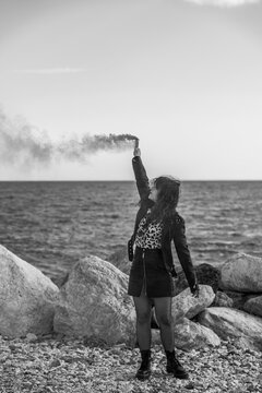black and white photo of an attractive woman in modern urban dress on the beach holding up a smoke flare with her arm.