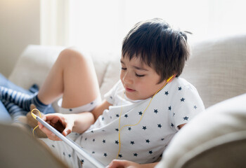 High key portrait kid wearing earphones listening to music, playing game on tablet,Boy sitting on sofa with light shining from window, Kid playing games on internet,Child doing homework online at home