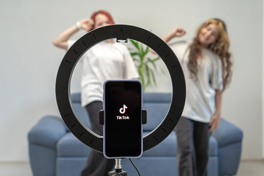 Two teenage girls are filming a video for their Tiktok vlog at their home. Tiktok logo on a phone on a tripod. Gen Z talent people play video selfie shoot app for show share viral story. 
