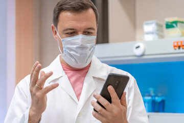 Excited doctor wearing white coat showing emotions of joy about victory in online lottery in room with laboratory box in UV ligh. Amazed male in medical mask win money celebration success