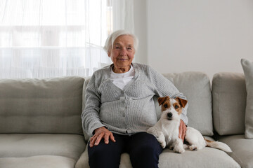 Emotional support animal concept. Portrait of elderly woman with wire haired jack russell terrier dog. Old lady and her rough coated pup sitting on grey textile sofa. Close up, copy space, background.