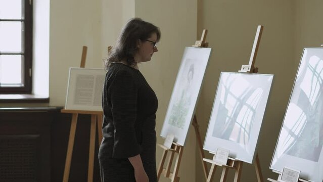 Young woman in black plaid dress, glasses examines paintings on easels at temporary art exhibition indoor medieval castle. Lady is culturally educated, carefully examines the artistic product.
