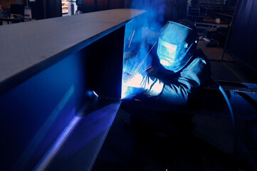 A welder in protective overalls and a welding mask welds metal structures by welding in a...