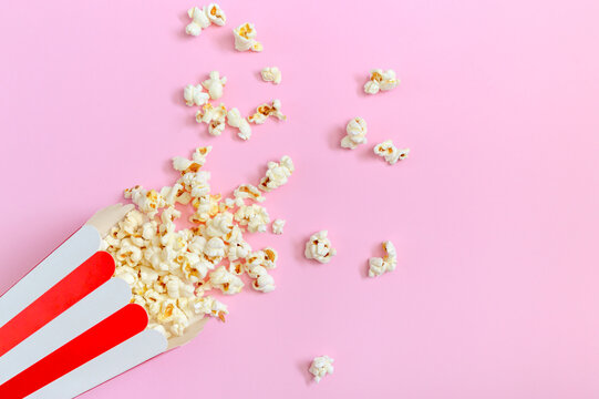 Popcorn spilled from a striped paper cup on a pink background with copy space. The concept of leisure, watching movies, shows, sports