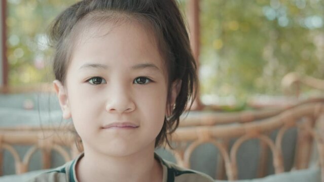 Face close up of adorable asian girl in comfortable living room at home looks at the camera with smiling and shows positive emotion, enjoyment, happiness of child with beautiful sunlight.